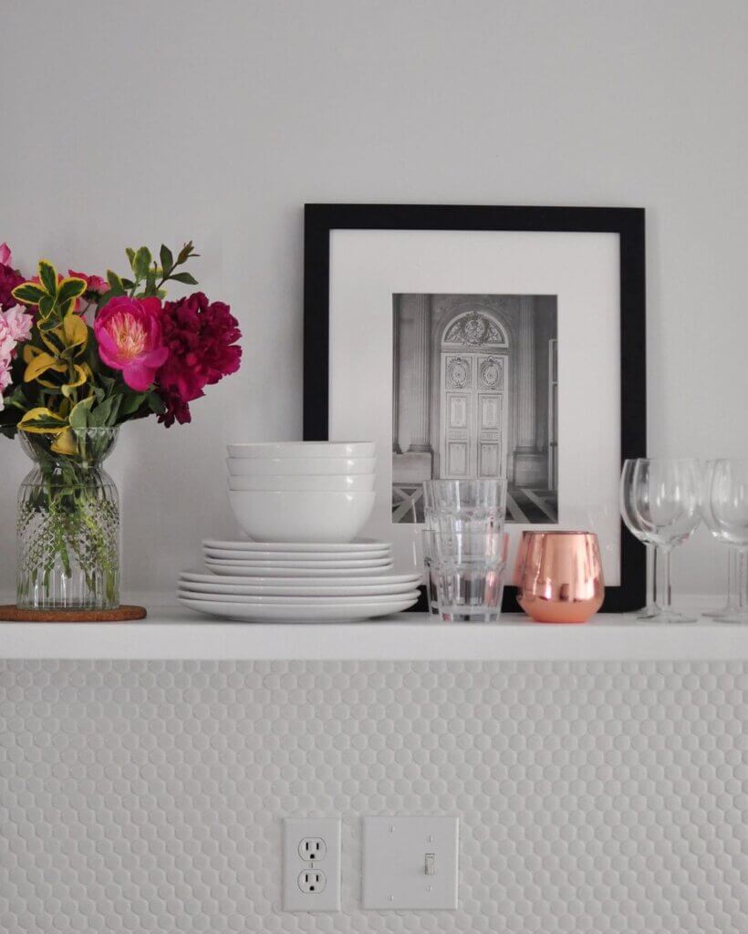 A black picture frame on a kitchen shelf with a vase of flowers and some glassware and dishes.